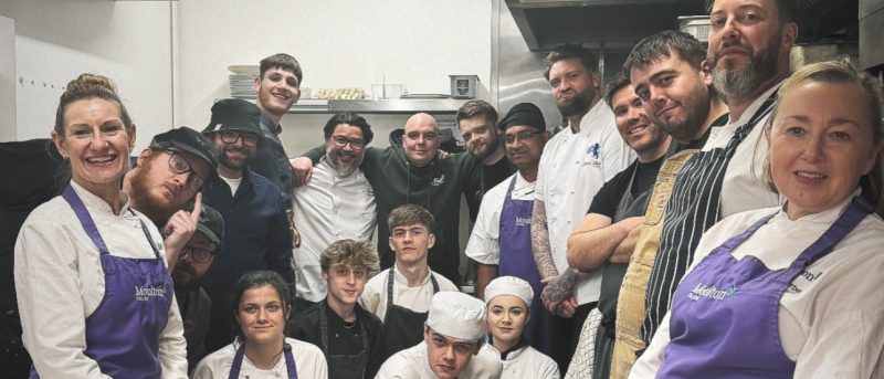 Group of chefs taking a group photo in the kitchen wearing purple moulton college aprons
