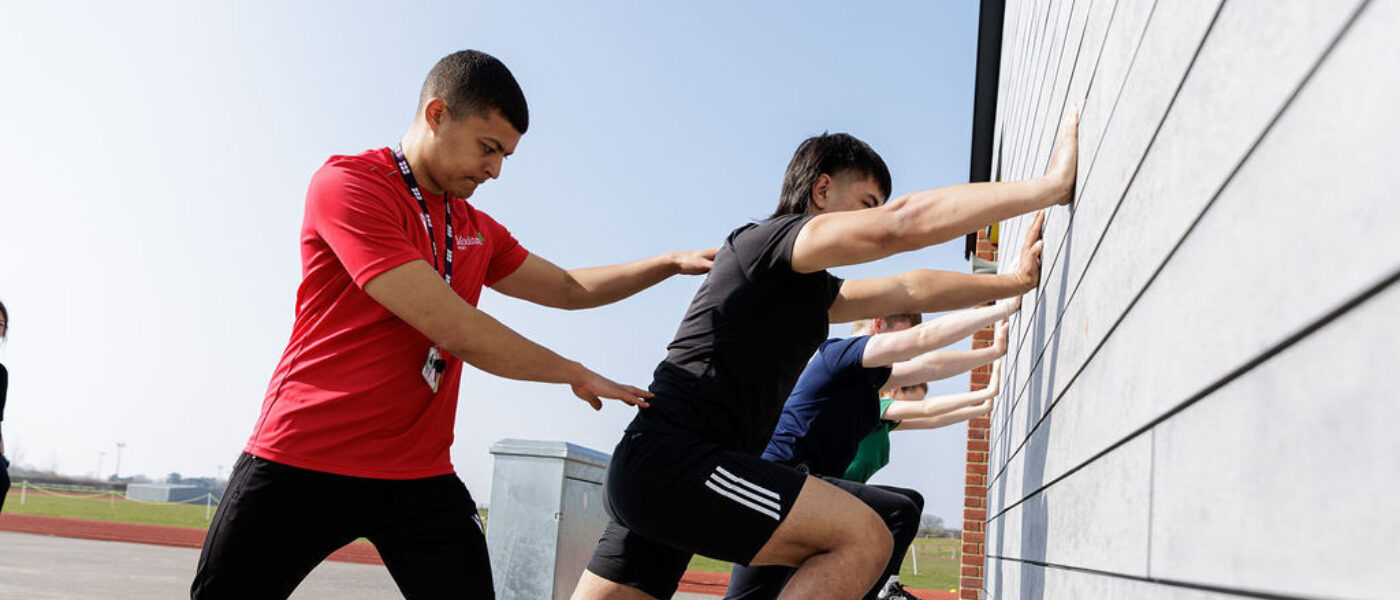 Students stretching against a wall directed by their tutor