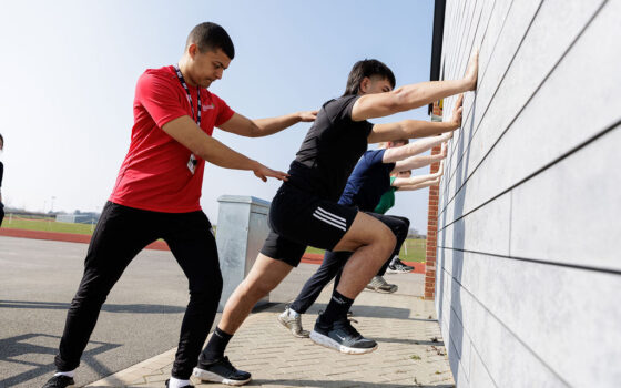 Students stretching against a wall directed by their tutor